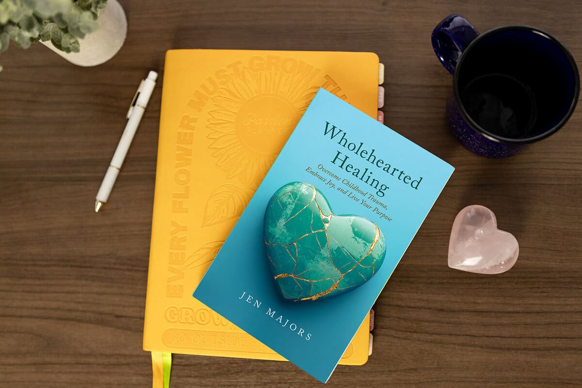 Wholehearted Healing paperback by Jen Majors on a yellow planner beside a coffee mug and a rose-quartz heart
